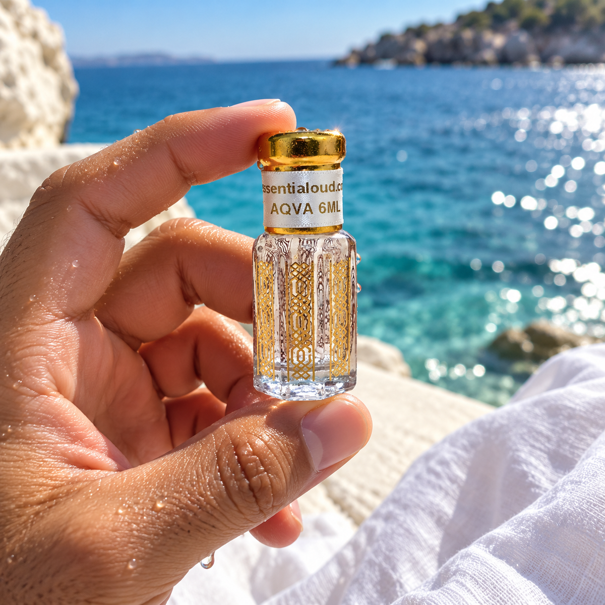 A hand holds a small clear glass perfume bottle with a gold cap and a white label reading 'Essential oud AQVA 6ML' against a sunny coastal background with blue sea and rocks.